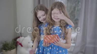 可爱的白种人黑发女孩闭上双胞胎妹妹的眼睛，给她打包的礼物。 感恩的孩子拥抱她可爱的