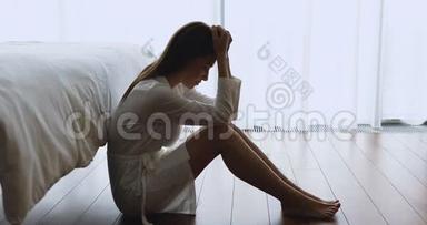 穿着睡衣坐在靠近床的地板上的年轻沮丧的女人。