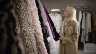 镜头接近年轻的金发白种人女人选择冬季大衣在精品店。 自信优雅的女士在