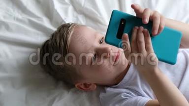 可爱的小男孩蹒跚学步，玩手机游戏，躺在床上拿着智能手机。 顶部视图