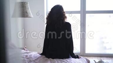 年轻的黑发女人坐在床上<strong>看</strong>着<strong>窗外</strong>。 穿着睡衣醒来的白种人女孩的后景