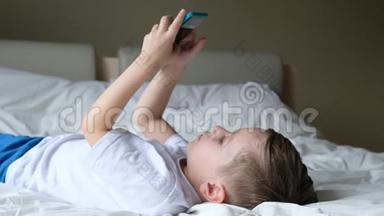 可爱的小男孩蹒跚学步，玩手机游戏，躺在床上，拿着智能手机