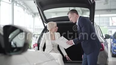 汽车经销商<strong>展示文件</strong>给年轻的金发白种人女人坐在汽车后备箱。 成功的商人