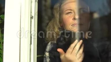 年轻的金发女人抱<strong>着</strong>一只猫，微笑<strong>着看着窗外</strong>。 科罗纳病毒检疫期间呆在家里