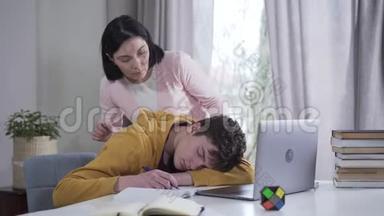 布鲁内特高加索人的母亲来到儿子<strong>身边</strong>，睡在桌子旁，叫醒他。 精疲力竭的十几岁男孩睡着了