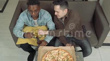 两个朋友在电视上看体育比赛。 男人在家坐在沙发上喝啤酒，吃披萨和零食