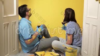 疲惫的一对男女坐在地板上，看着油漆过的黄墙，喝着酒，他们累了，但却累了
