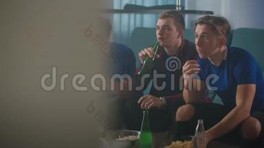 三个穿着蓝色T恤的好朋友坐在沙发上喝啤酒，看电视时跳着体育节目