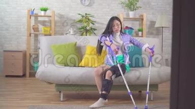 一位年轻的亚洲妇女受伤后，膝盖弯曲，膝盖支撑，用拐杖从沙发上站起来