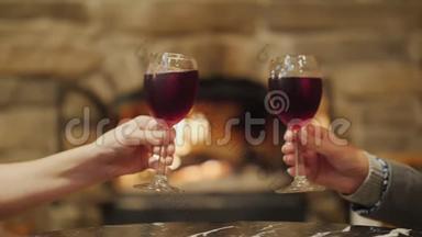 两只手拿着一杯酒，在一个燃烧的壁炉的背景下叮当作响。 家里的节日和温馨