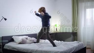 活跃的小红发男孩在床上跳跃，听音乐。 快乐的白种人孩子周末在家玩