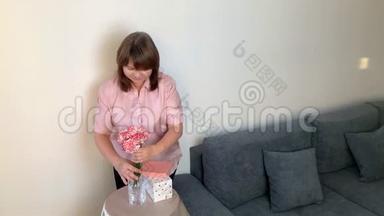 女人把一束粉红色的康乃馨放在花瓶里，微笑着，盒子里的礼物放在<strong>圆桌</strong>上，节日的概念，情人节`