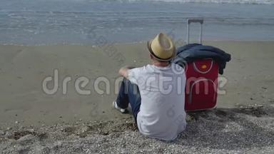 旅行者戴着帽子坐在海边的沙滩上，旁边是一个红色的手提箱，看着海浪