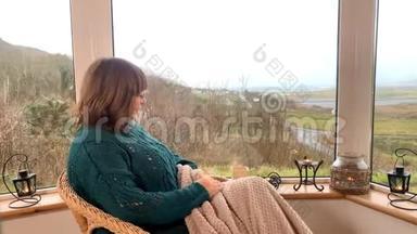 女人坐在一张铺着格子的舒适的柳条椅子上，坐在山上一个灯光封闭的阳台上，喝着热茶，<strong>看</strong>着<strong>窗外</strong>，