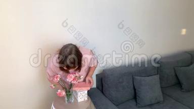 女人闻着一束粉红色的康乃馨，微笑，盒子里的礼物躺在圆桌上
