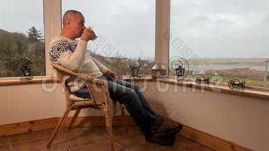 男人坐在阳台上舒适的柳条椅子上，喝着热茶，<strong>望</strong>着<strong>窗外</strong>，享受和享受的概念