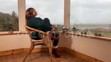 女人坐在一张舒适的柳条椅上，坐在山上一个灯光封闭的阳台上，享受着放松，看着窗外，紧紧抓住她