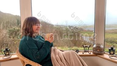 女人坐在阳台窗边舒适的柳条椅子上，喝着热茶，躲在毯子里，看着窗外，一个