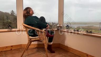 女人坐在一张舒适的柳条椅上，坐在山上一个灯光封闭的阳台上，享受着放松，<strong>看</strong>着<strong>窗外</strong>，紧紧抓住她