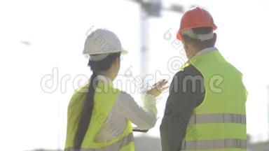 一名男子和一名年轻女子在一个建筑工地戴着头盔和信号背心。 女人向她汇报一些事情