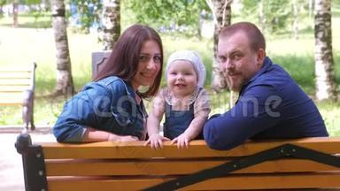 一家人妈妈，宝贝女儿和爸爸坐在公园的长凳上，微笑着看着摄像机。
