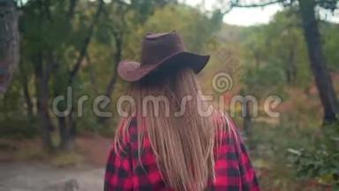 一个长发女孩穿着一件黑色和红色的衬衫，戴着一顶牛仔帽，正穿过树林。