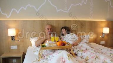微笑的女人和男人躺在酒店房间的床上，在一张桌子前，集中着水果和饮料，早餐在床上。