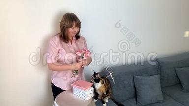 女人捧着一束粉红色的康乃馨花，看着尝试，盒子里的礼物放在圆桌上，一只黑猫坐在圆桌上