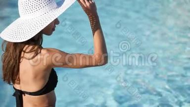 特写镜头。 模糊的水背景。 一个戴着白色大帽子的年轻女人坐在游泳池附近，戴着她的帽子。 有地方住