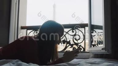 快乐美丽的欧洲女人在智能手机应用程序上输入信息，躺在阳台床上，埃菲尔铁塔观景慢