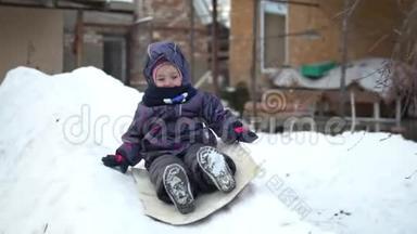 一个孩子在雪地滑梯上翻滚。 冬天在街上玩。 在寒冷的冬天，孩子穿着一件夹克和一件