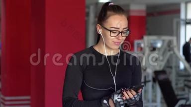 带着耳机的年轻女孩在健身房里用智能手机。 女孩听音乐