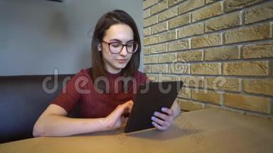 一个拿着平板电脑的年轻女人坐在桌子旁。 一个女人坐在家里的一张桌子上，桌子上有一块平板电脑，正在通信。