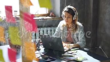 有创意的年轻女商人正在用耳机听音乐，在办公桌前用笔记本电脑跳舞