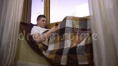 一个年轻人正在打电话。 一个男人躺在窗边的窗台上，手里拿着一部智能手机。 外部