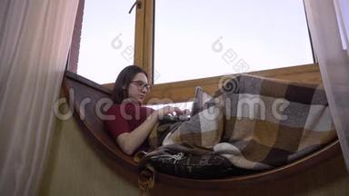 一个年轻的女人正在笔记本电脑上打字。 一个女孩躺在窗边的窗台上，手里拿着一台笔记本电脑。 在外面
