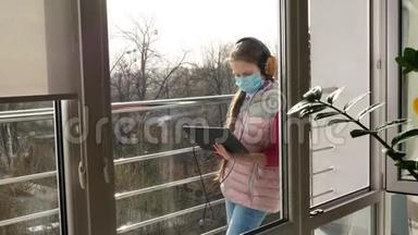 十几岁的女孩戴着面具和耳机，打字，在平板电脑上工作，在开放的阳台上。 春天阳光明媚的一天。 远程学习