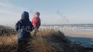 小女孩和男孩穿着温暖的外套，在阳光明媚的冬天，海滩缓慢的运动中，沿着高大的草地沙丘行走。