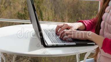 手在笔记本电脑键盘上打字的特写。 青少年女孩打字，在笔记本电脑上工作，在开放的阳台上。 春天阳光明媚