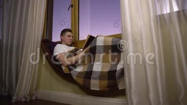 一个年轻人正在看书。 一个人躺在窗边的窗台上，手里拿着一本书。 晚上出去