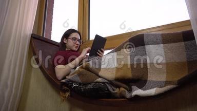 一位年轻女子正在平板电脑上发短信。 一个女孩躺在窗边的窗台上，手里拿着一块平板电脑。 外部