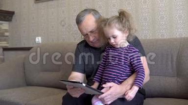 孙女和祖父坐着一块石碑。 女孩对平板皱着眉头。 一起坐在沙发上。