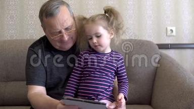 孙女和祖父坐着一块石碑。 女孩对平板皱着眉头。 一起坐在沙发上。