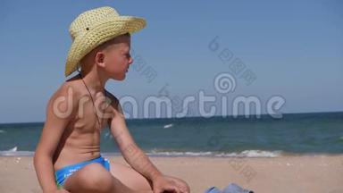 男孩坐在金沙上。 这孩子戴着一顶大<strong>帽檐</strong>的草帽. 孩子在沙滩上玩