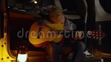 小男孩吉他手在森林徒步旅行中在汽车后备箱上弹吉他。 带游客玩吉他音乐的少年男孩