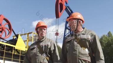两名戴着橙色头盔的石油工人在油泵千斤顶附近行走和交谈。 石油<strong>工程师</strong>监督原油<strong>现场</strong>