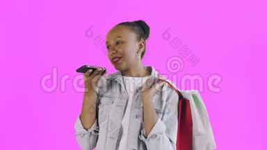 美国黑人妇女带着购物袋的肖像正在粉色背景上通过智能手机录制一条语音信息。