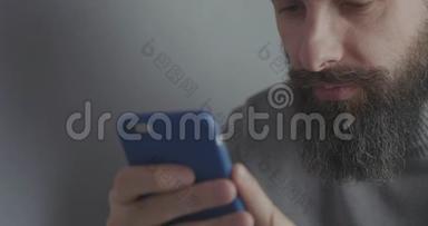 漂亮的大胡子成年男子在手机屏幕上观看，通过社交媒体使用智能手机滚动。 快乐的特写
