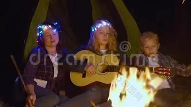 十几岁的女孩在森林远足中弹吉他，唱着篝火前的歌。 友好的家庭在露营时唱歌
