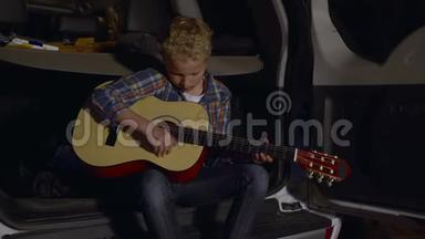 坐在汽车后备箱里用吉他<strong>演奏</strong>音乐的少年男孩。 旅游男孩吉他手在夏令营中<strong>演奏</strong>音乐